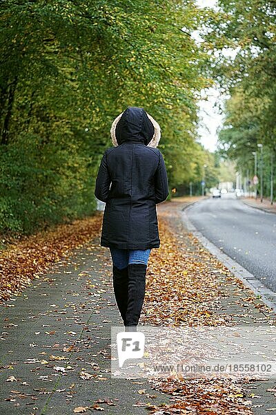 Rückansicht einer nicht erkennbaren Frau in Wintermantel und Stiefeln  die im Herbst die Straße entlanggeht