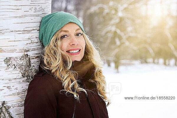 Lächelnde Frau lehnt sich gegen eine Birke und genießt den sonnigen Tag in einer schneebedeckten Winterlandschaft  mit Sonnenlichtleck