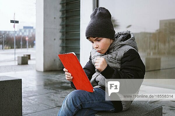 Aufgeregter kleiner Junge  der ein Online-Spiel auf einem Tablet-Computer spielt  während er draußen allein auf einer Stadtstraße im Winter sitzt und seinen Sieg mit geballter Faust feiert