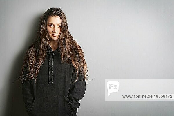 cool young woman with long brunette hair wearing black hoodie sweater leaning against wall with her hands in pockets - gray background with copy space