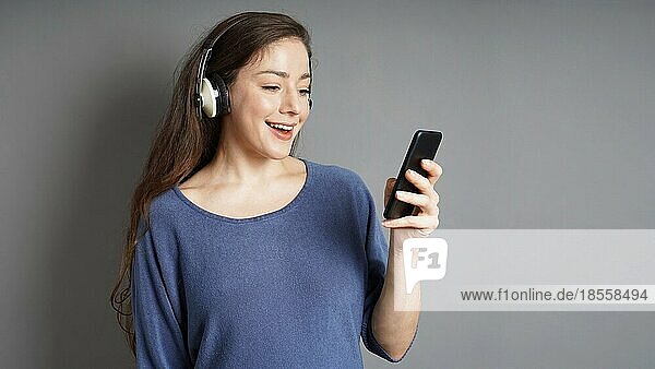 Glückliche junge Frau mit Kopfhörern  die Musik auf ihrem Smartphone streamt - grauer Hintergrund mit Kopierraum