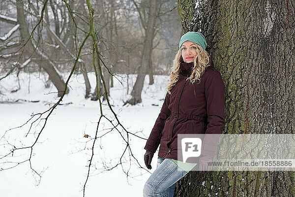 Blonde Frau lehnt an einem Baum in einem verschneiten Park im Winter