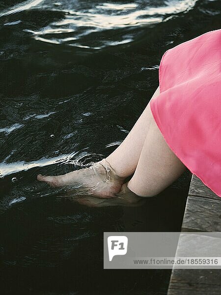 Unerkennbares Mädchen junge Frau sitzt auf Steg am See und taucht nackte Füße ins Wasser