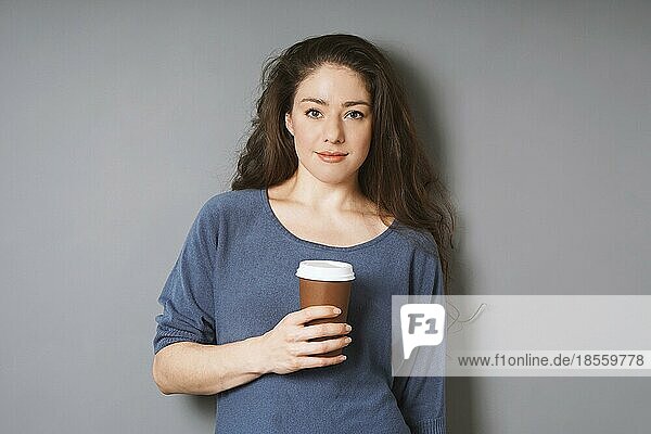 relaxed young woman on her coffee break - holding coffee to go in disposable paper cup while leaning against gray wall