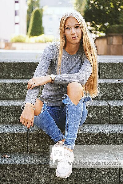 Teenager-Mädchen mit langen blonden Haaren und ausgebeulten Jeans sitzt draußen auf den Stufen - urbaner Lebensstil oder Straßenmode-Konzept