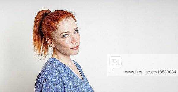 Junge Frau mit roten Haaren  die zu einem Pferdeschwanz gebunden sind  neigt ihren Kopf und lächelt auf neutralem Hintergrund mit Leerzeichen