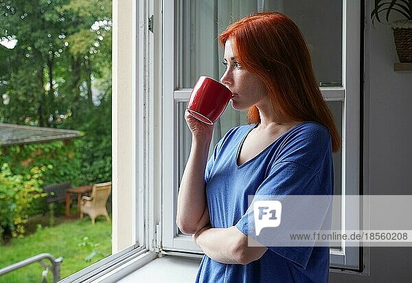 young woman standing by open window drinking from coffee cup while looking out into garden