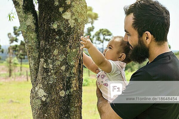 Niedliches Baby  das sich im Frühlingswald an einen Baumstamm schmiegt  sein Vater hält es in seinen Armen