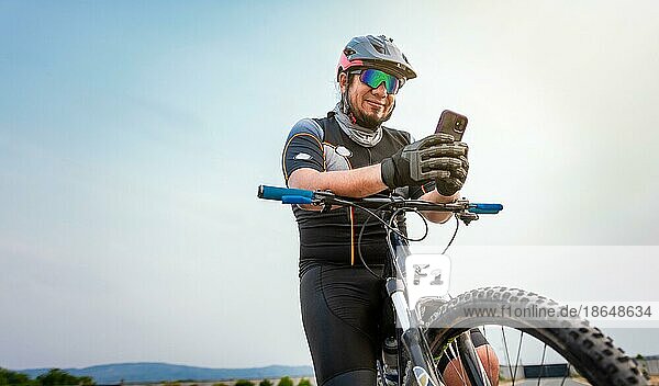 Männlicher Radfahrer mit Helm und Mobiltelefon. Radfahrer auf seinem Fahrrad SMS mit Telefon im Freien. Radfahrer Männchen mit Helm und Brille SMS auf Handy im Freien