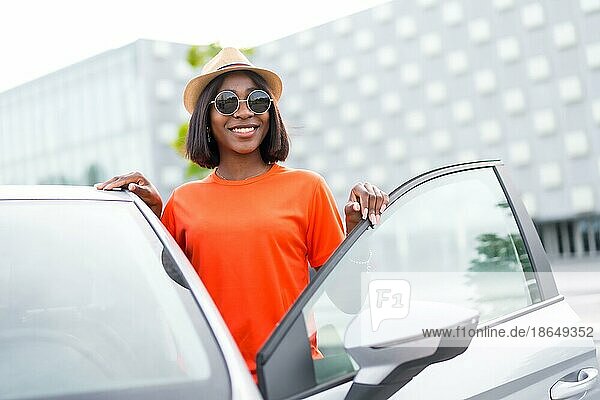 Stilvoller Sommer: schwarze Frau in orangefarbenem Hemd und Sonnenbrille öffnet lächelnd die Autotür