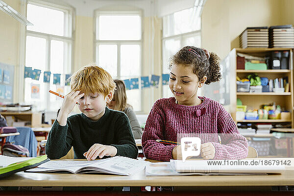 Smiling girl sitting by confused blond boy at desk in classroom