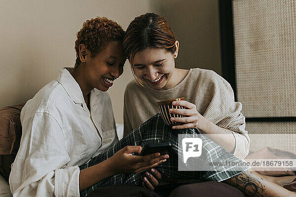 Glückliches nicht-binäres Paar  das zu Hause auf dem Bett sitzt und auf sein Smartphone schaut