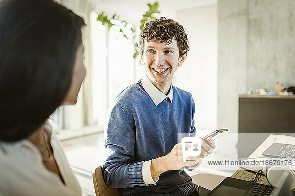 Happy young entrepreneur holding smart phone discussing with colleague in creative office
