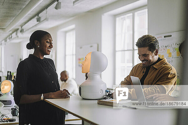Multirassische lächelnde Studenten mit beleuchtetem sozialen Roboter am Schreibtisch im Innovationslabor