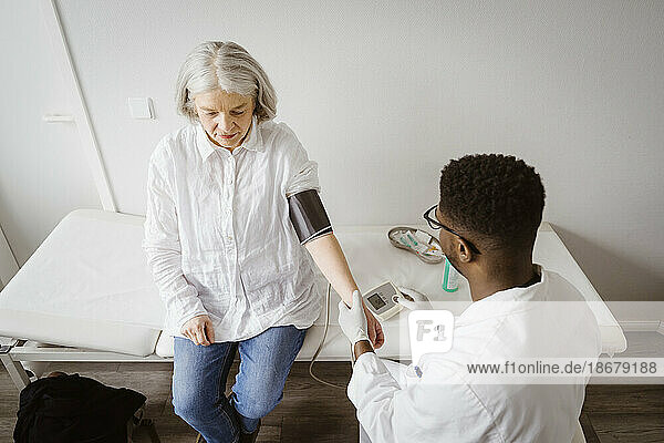 High angle view of male doctor measuring blood pressure of senior patient sitting on bed at medical clinic