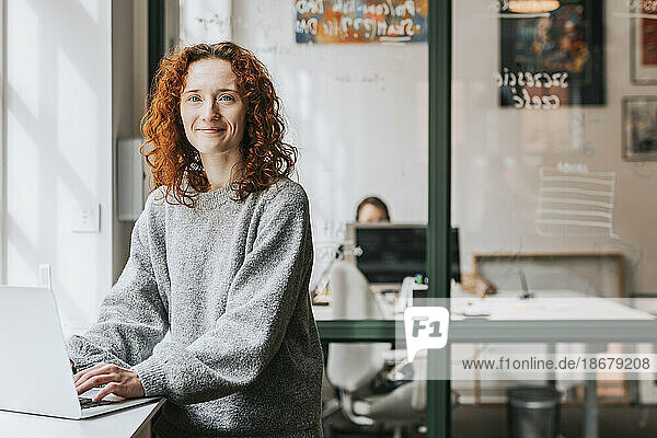 Porträt einer lächelnden rothaarigen Geschäftsfrau mit Laptop auf der Fensterbank im Büro