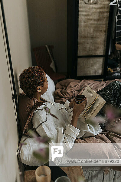 Hoher Blickwinkel einer nicht-binären Person  die ein Buch liest  während sie zu Hause auf dem Bett sitzt