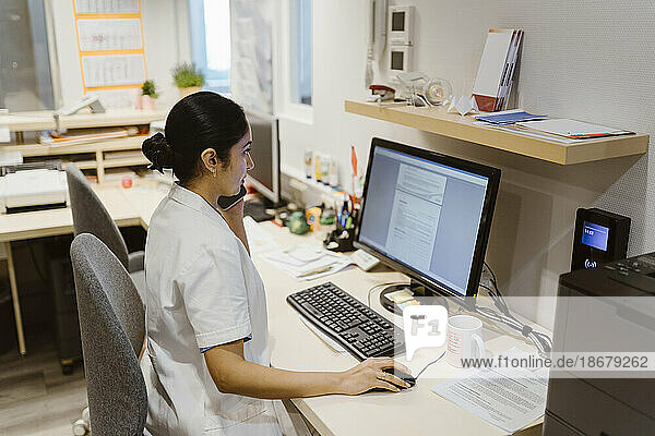 Mitarbeiterin des Gesundheitswesens benutzt einen Computer  während sie in einer Klinik mit einem Smartphone telefoniert
