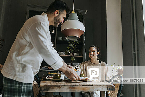 Lächelnder Mann  der das Frühstück mit einer nicht-binären Person serviert  die am Esstisch in einem Haus sitzt