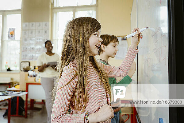 Happy girl writing on whiteboard while solving maths problem in classroom