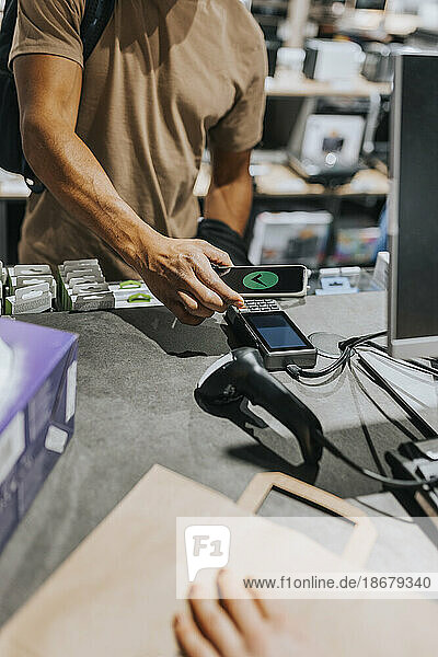 Mittelteil eines männlichen Kunden  der ein Kreditkartenlesegerät über ein Smartphone an der Kasse eines Elektronikgeschäfts scannt