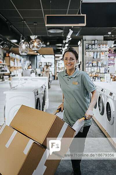 Porträt einer lächelnden Verkäuferin  die in einem Elektronikgeschäft Kisten im Einkaufswagen transportiert