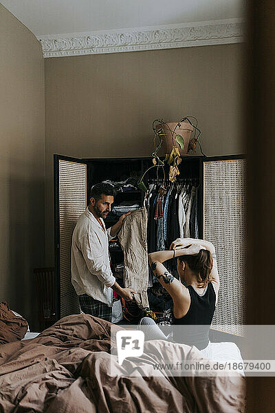 Mann zeigt Kleid an nicht-binäre Person  die sich die Haare bindet  während sie zu Hause auf dem Bett sitzt