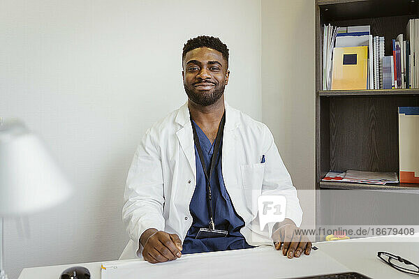Porträt eines lächelnden männlichen Mitarbeiters im Gesundheitswesen  der einen Laborkittel trägt und am Schreibtisch in einer Klinik sitzt