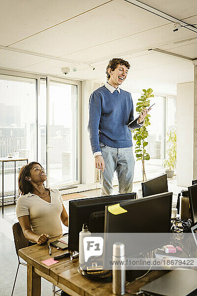 Glücklicher Unternehmer auf einem Stuhl stehend neben einem Kollegen am Schreibtisch in einem kreativen Büro