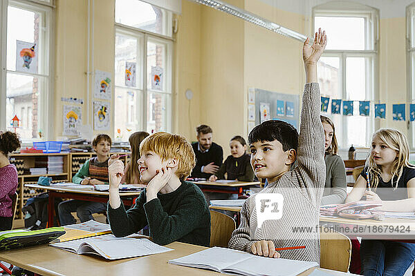 Boy with hand raised answering during lecture while sitting by male friend in classroom