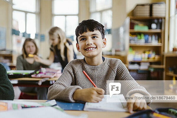 Lächelnder männlicher Schüler  der einen Bleistift hält  während er am Schreibtisch im Klassenzimmer sitzt