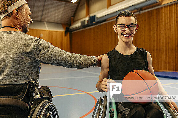 Männlicher Sportler mit Behinderung motiviert ein Mädchen  das im Rollstuhl sitzt  mit einem Basketball auf dem Sportplatz