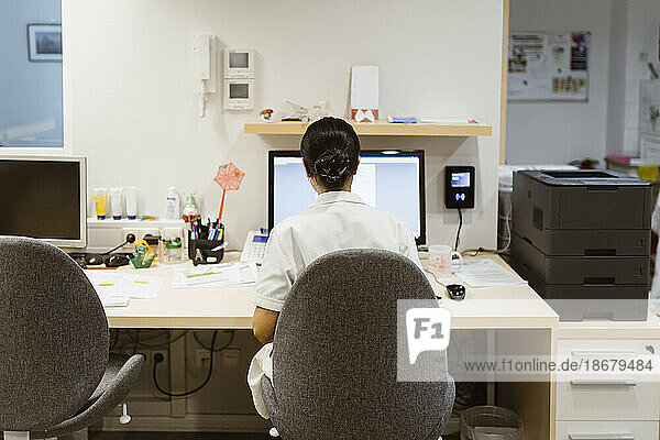 Rückansicht einer Ärztin  die in einer Klinik auf einem Stuhl sitzend an einem Desktop-Computer arbeitet
