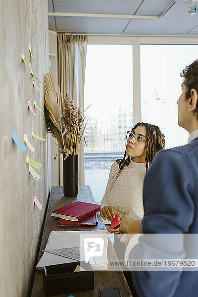 Male and female colleagues discussing over sticky notes on wall in creative office