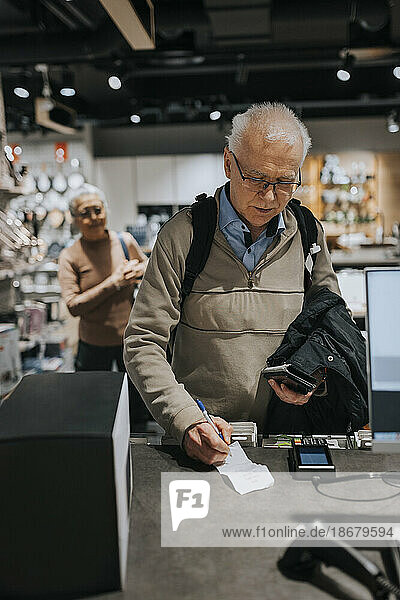 Ein älterer Mann unterschreibt eine Rechnung mit einem Kreditkartenlesegerät an der Kasse eines Haushaltswarengeschäfts