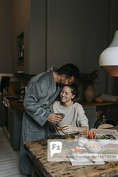 Mann küsst lächelnde nicht-binäre Person beim Frühstück zu Hause