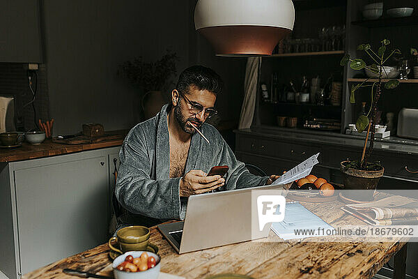 Mann hält Rechnungen und benutzt ein Smartphone  während er mit einem Laptop auf dem Esstisch zu Hause sitzt