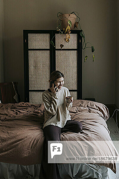 Lächelnde nicht-binäre Person  die über ihr Mobiltelefon spricht und zu Hause auf dem Bett sitzt