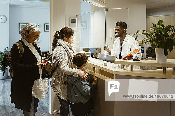 Patients standing and talking to male receptionist through transparent shield in clinic