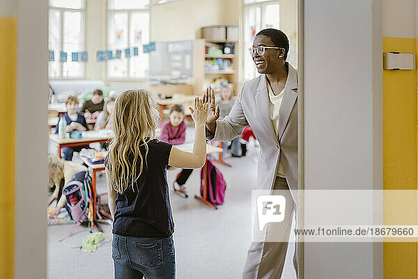 Smiling female teacher and schoolgirl giving high-five at doorway in classroom