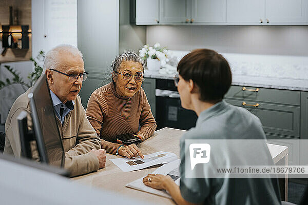 Ein älteres Paar diskutiert mit einem Innenarchitekten über eine Broschüre  während es am Schreibtisch in einem Geschäft sitzt.