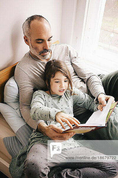 Vater und Tochter lesen ein Buch  während sie zusammen auf einer Fensternische zu Hause sitzen