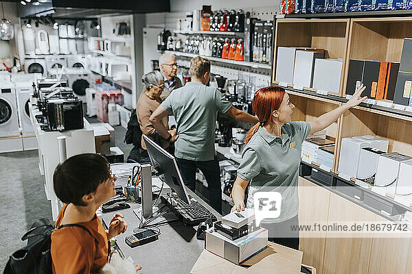Female sales clerk showing various appliances on shelf to customer at checkout counter