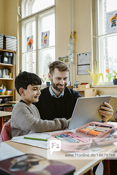 Happy male teacher assisting schoolboy using tablet PC at desk in school