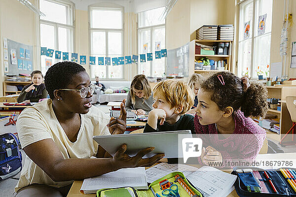 Lehrerin mit Tablet-PC  die einem Schüler und einer Schülerin am Schreibtisch erklärt  während sie im Klassenzimmer gestikuliert