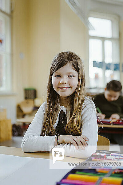 Portrait of smiling schoolgirl sitting with arms crossed at desk in classroom