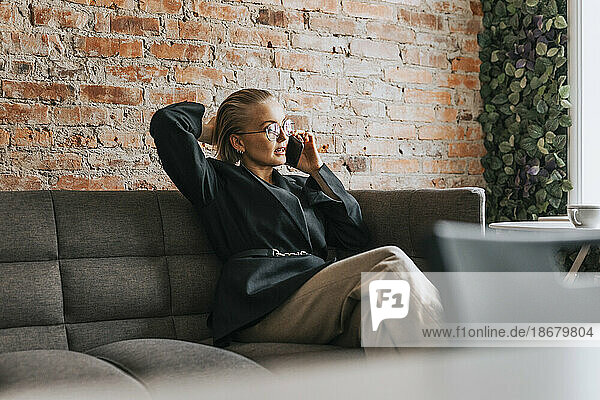 Junge Geschäftsfrau mit Hand im Haar  die auf einem Sofa im Büro sitzt und mit ihrem Smartphone spricht