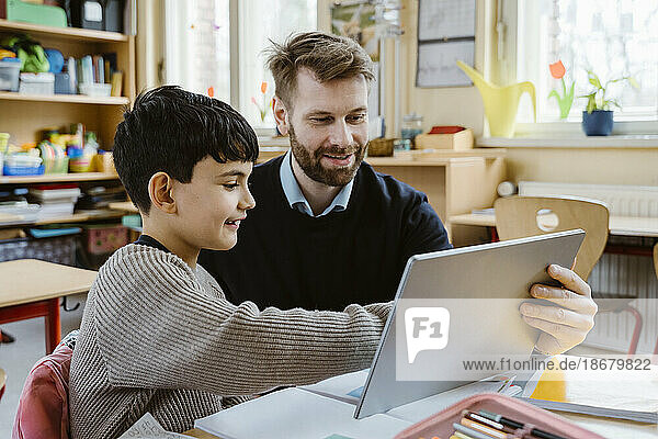 Lächelnder männlicher Lehrer  der einem Schüler bei der Benutzung eines Tablet-PCs am Schreibtisch in der Schule hilft
