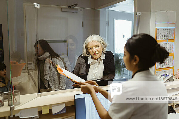Ältere Patientin  die das Ausfüllen des Formulars durch die Empfangsdame durch eine transparente Scheibe in der Klinik versteht