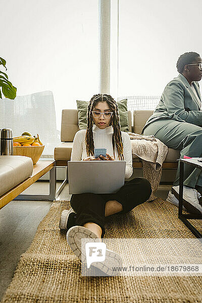 Weiblicher Programmierer  der ein Smartphone benutzt  während er mit seinem Laptop auf dem Teppich im Büro sitzt